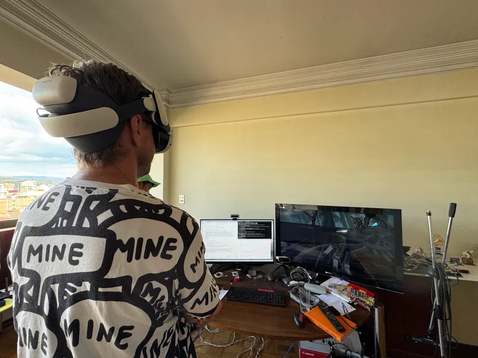 Una persona con un visor de realidad virtual está de pie en un cuarto con un escritorio, monitores y una ventana hacia la ciudad. Vista de espaldas.