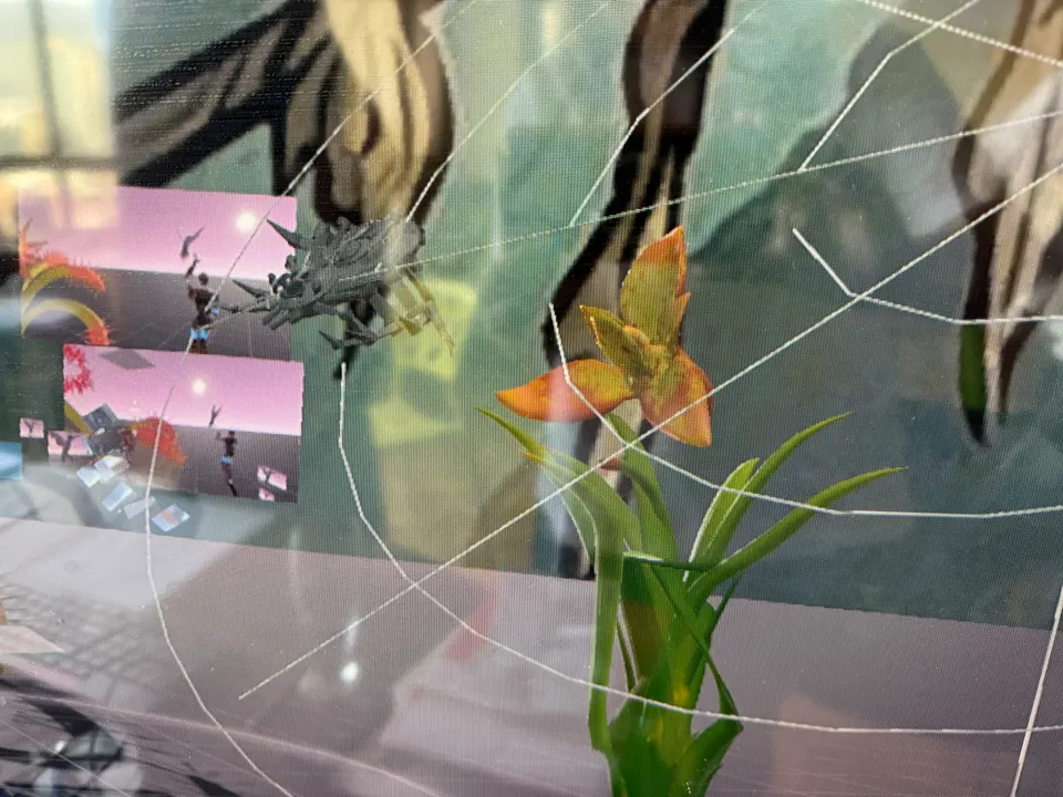 Una araña 3D en su tela junto a una flor naranja. Dos ventanas pequeñas muestran un mundo virtual rosado.