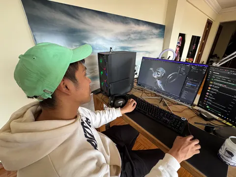 Una persona con gorra verde trabaja en un escritorio con dos monitores. Uno muestra una criatura 3D en Blender.
